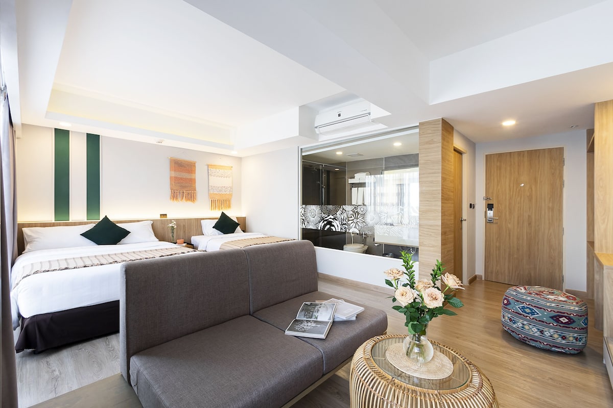 Una habitación de hotel moderna con una cama grande, un sofá, una pequeña mesa redonda con un jarrón de flores y una puerta de madera.