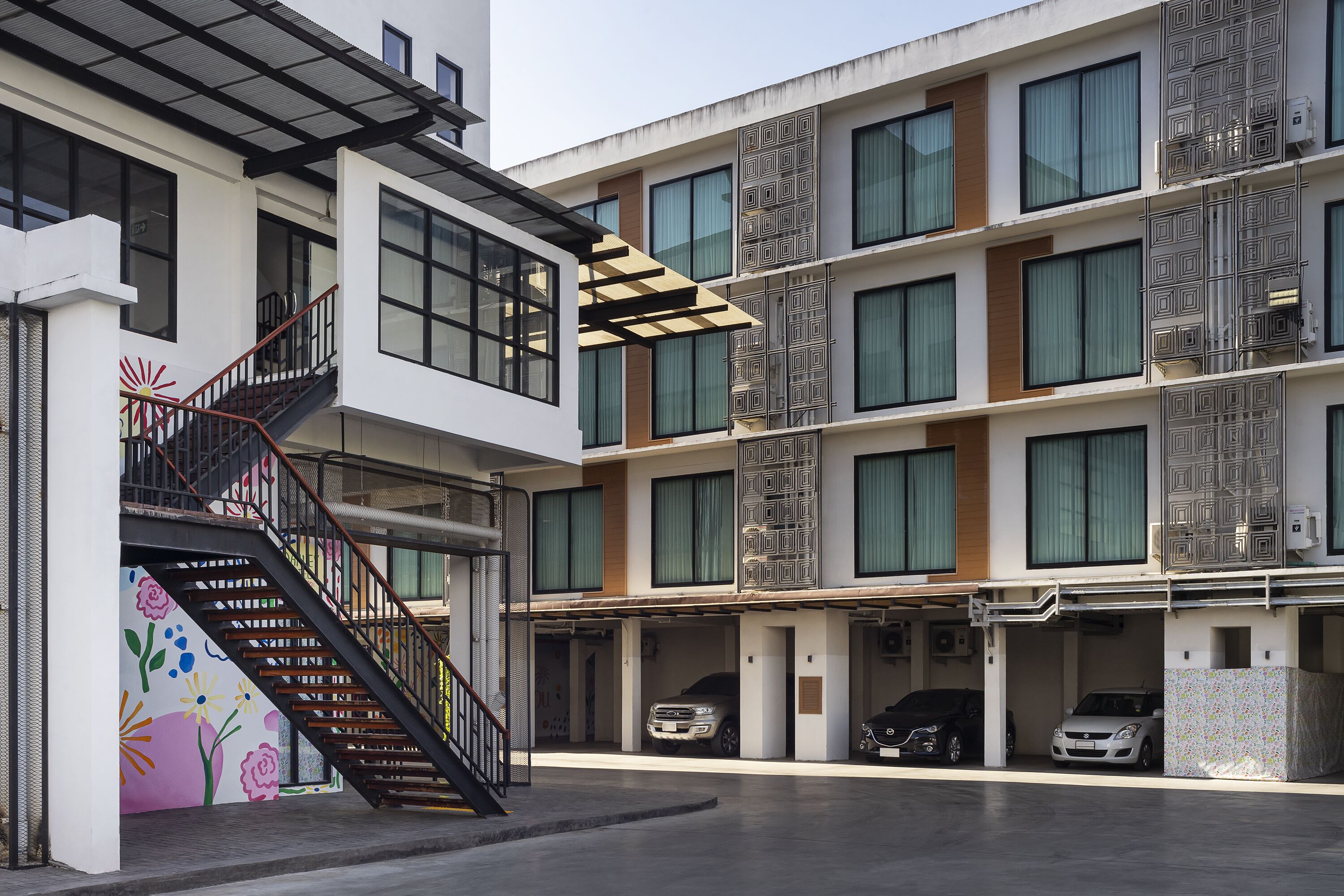 A multi-story building with a modern design, featuring a staircase with a metal railing, large windows, and a parking area with several cars.