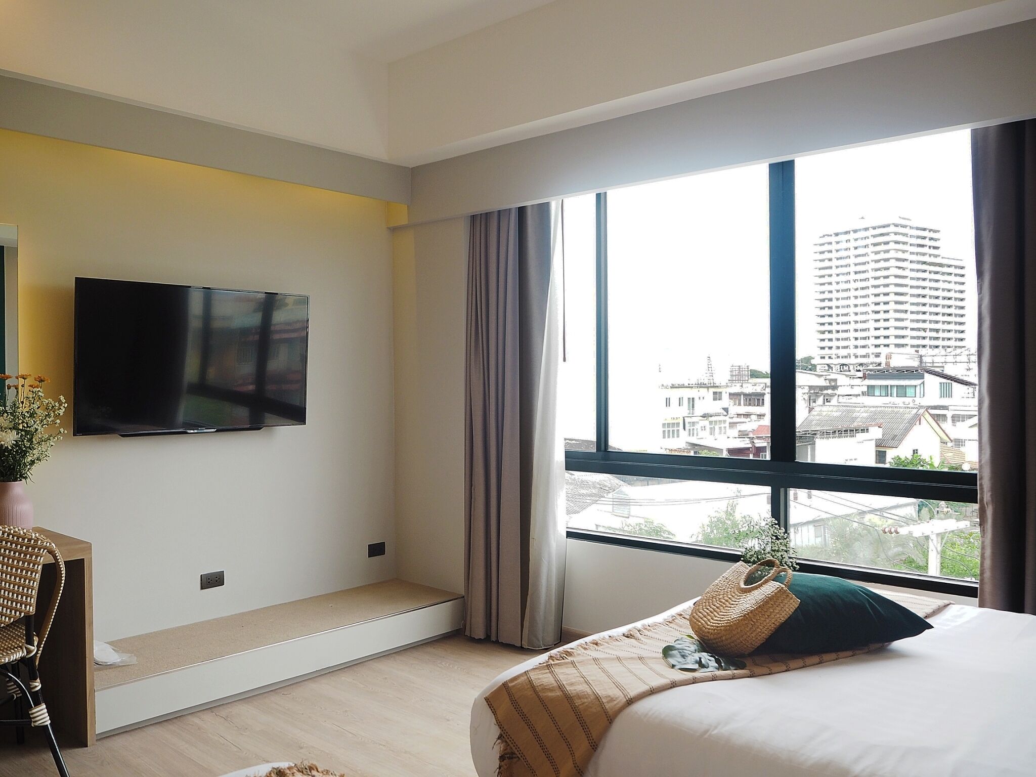Una habitación de hotel moderna con un ventanal grande, un televisor de pantalla plana en la pared, una cama con un cojín verde y una silla de mimbre.