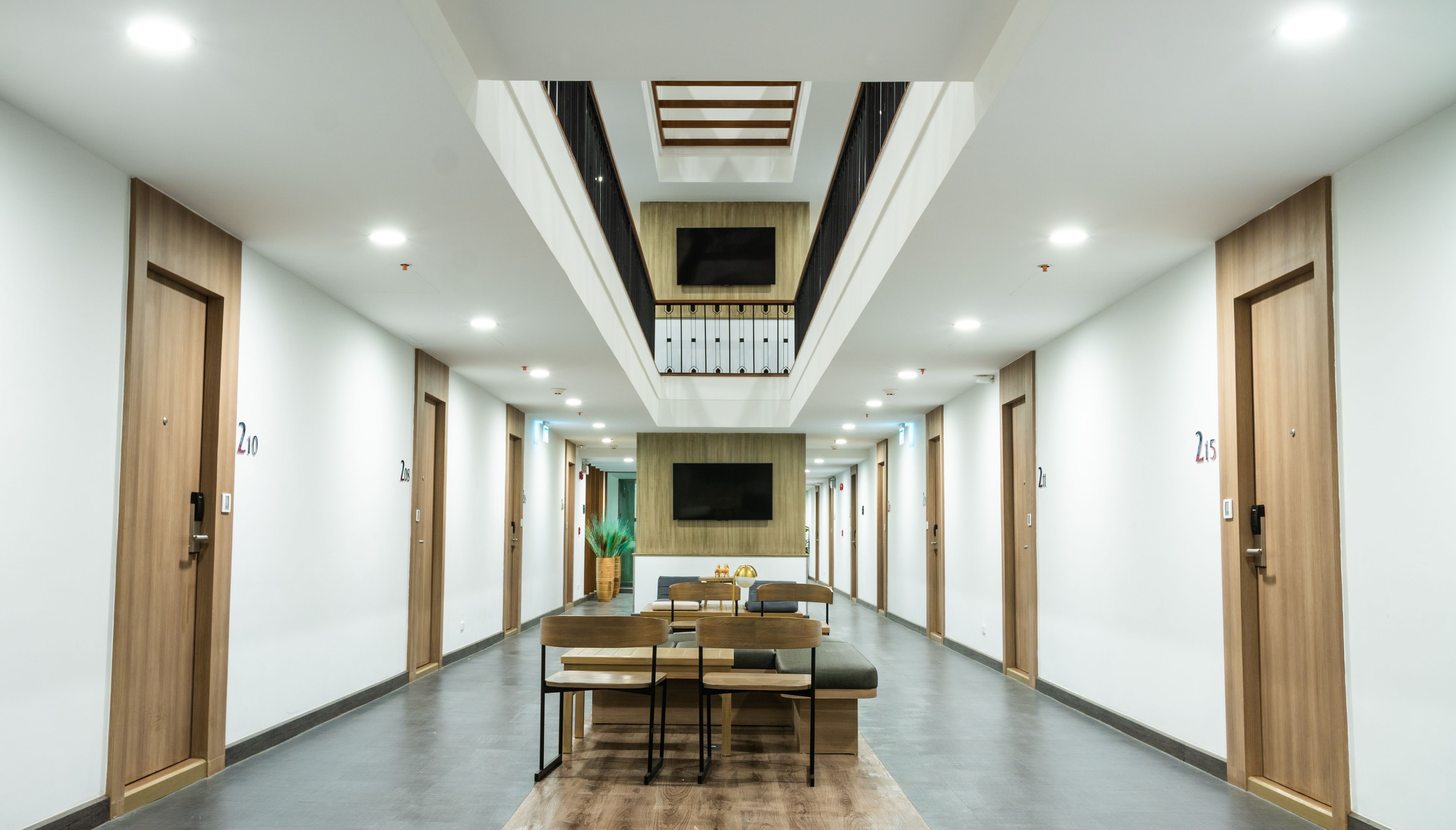 Ein Hotelkorridor mit Holztüren, einer zentralen Sitzecke mit Tischen und Stühlen sowie einem an der Wand befestigten Flachbildfernseher.