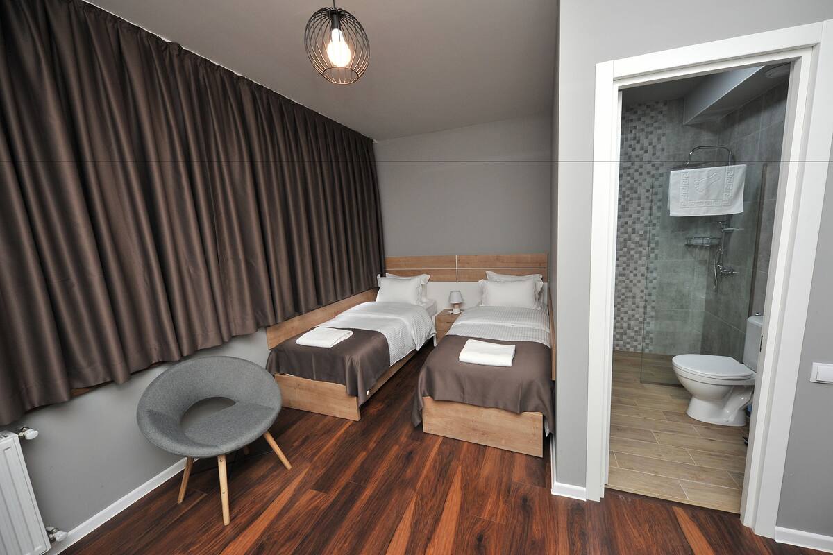 A modern hotel room with two beds, a grey chair, a wooden floor, and a bathroom visible through an open door.
