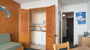 Een compacte kamer met houten vloer, een groene bank, een houten eettafel en een keuken met witte kastjes en een betegelde achterwand.
