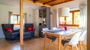 A modern living room with a dining area, wooden beams, a cabinet, and a window with curtains. - Alpenchalet Sachrang für 2 Personen (Sachrang)