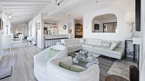 A spacious living room with a large sectional sofa, a glass coffee table, and a dining area with a kitchen in the background. - H242: Modern 1BD Villa, Fantastic Ocean View, Fireplace, Golf Cart (Avalon)