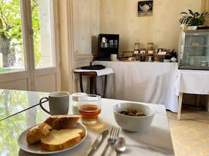 Une table de petit-déjeuner avec du pain, un bol de céréales, une tasse et un verre de jus. On aperçoit, à l’arrière-plan, un meuble contenant des ustensiles de cuisine et une plante.