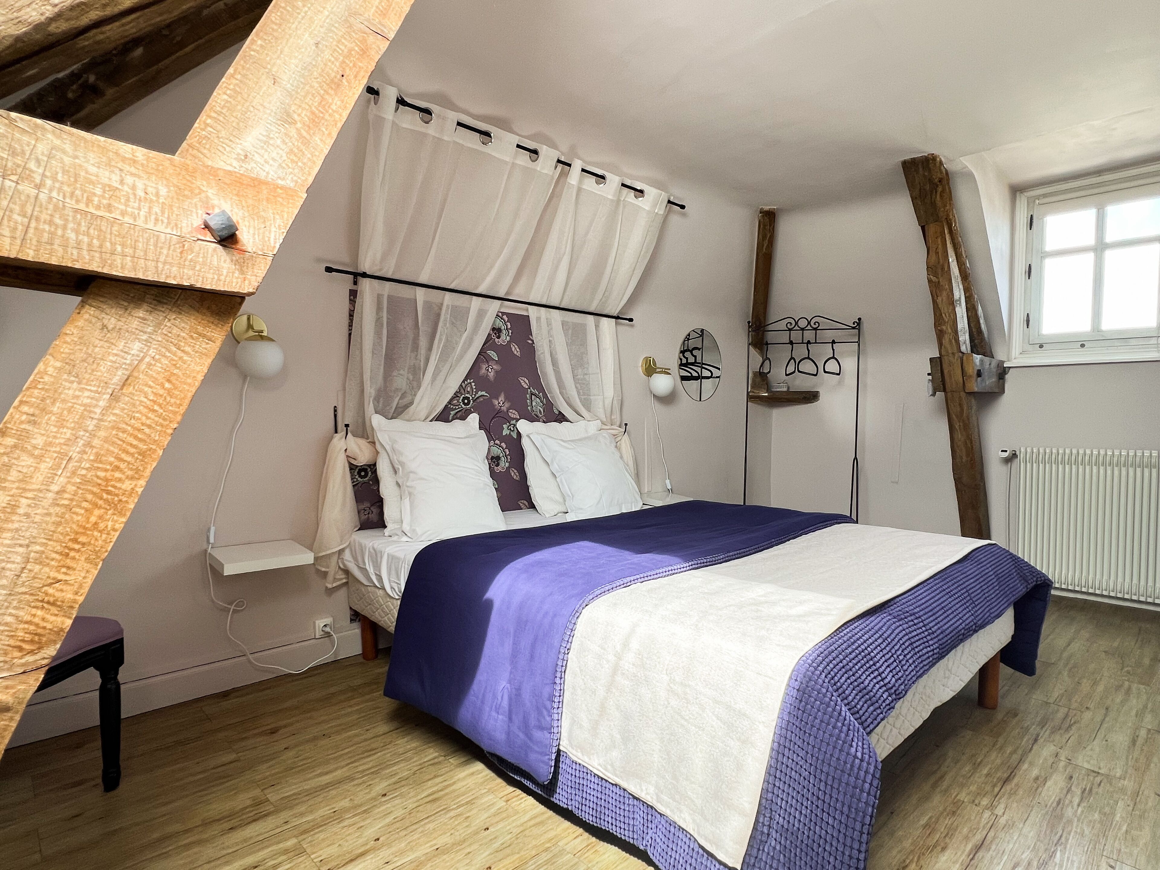 A bedroom with a bed, a window, a radiator, and a wooden beam structure.