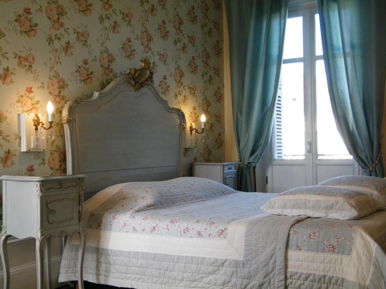A bedroom with a bed, nightstands, a dresser, floral wallpaper, and a window with curtains.