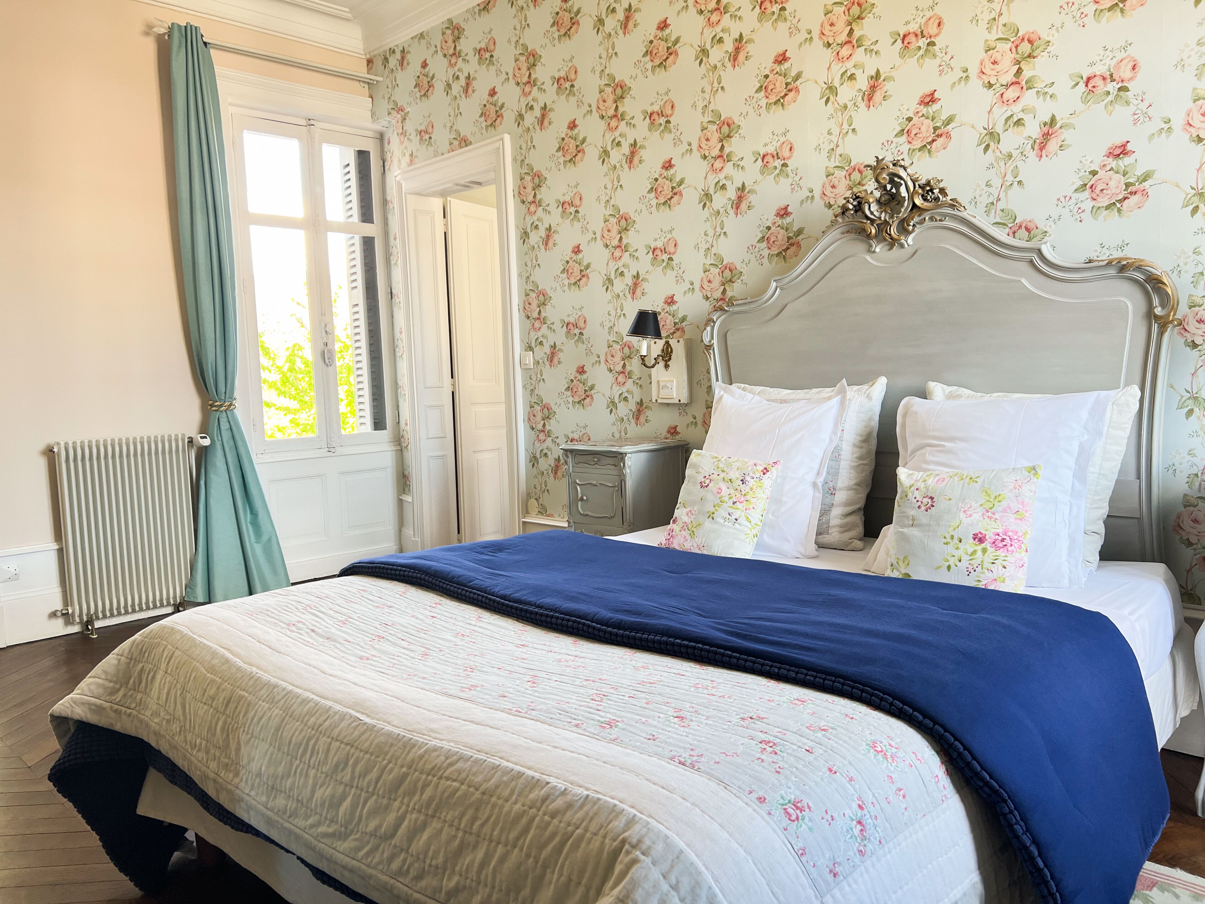 A bedroom with a bed, floral wallpaper, a radiator, and a window with curtains.