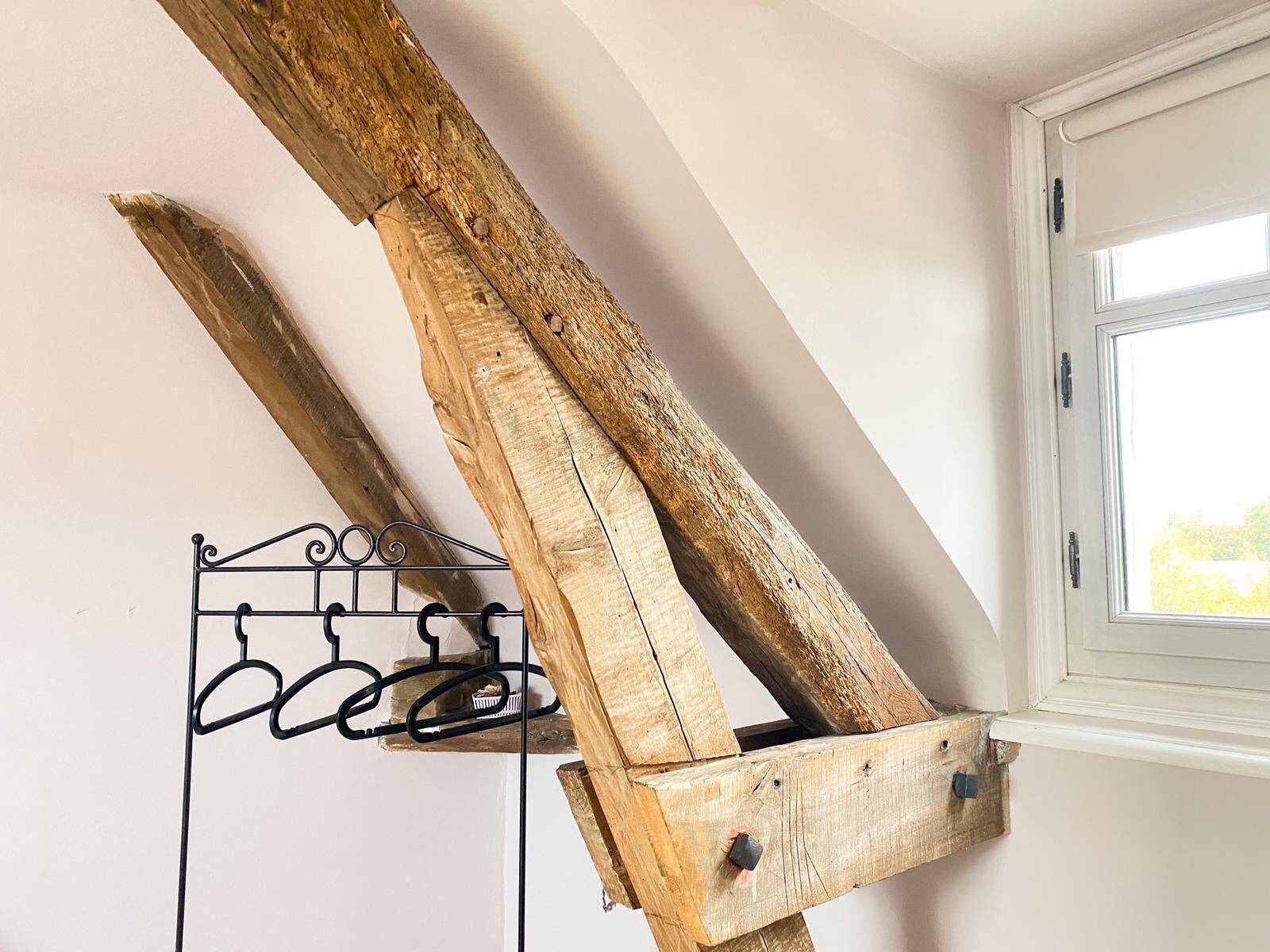 Un perchero de madera rústico con sistema de ganchos metálicos sobre una pared blanca, cerca de una ventana con marco blanco.