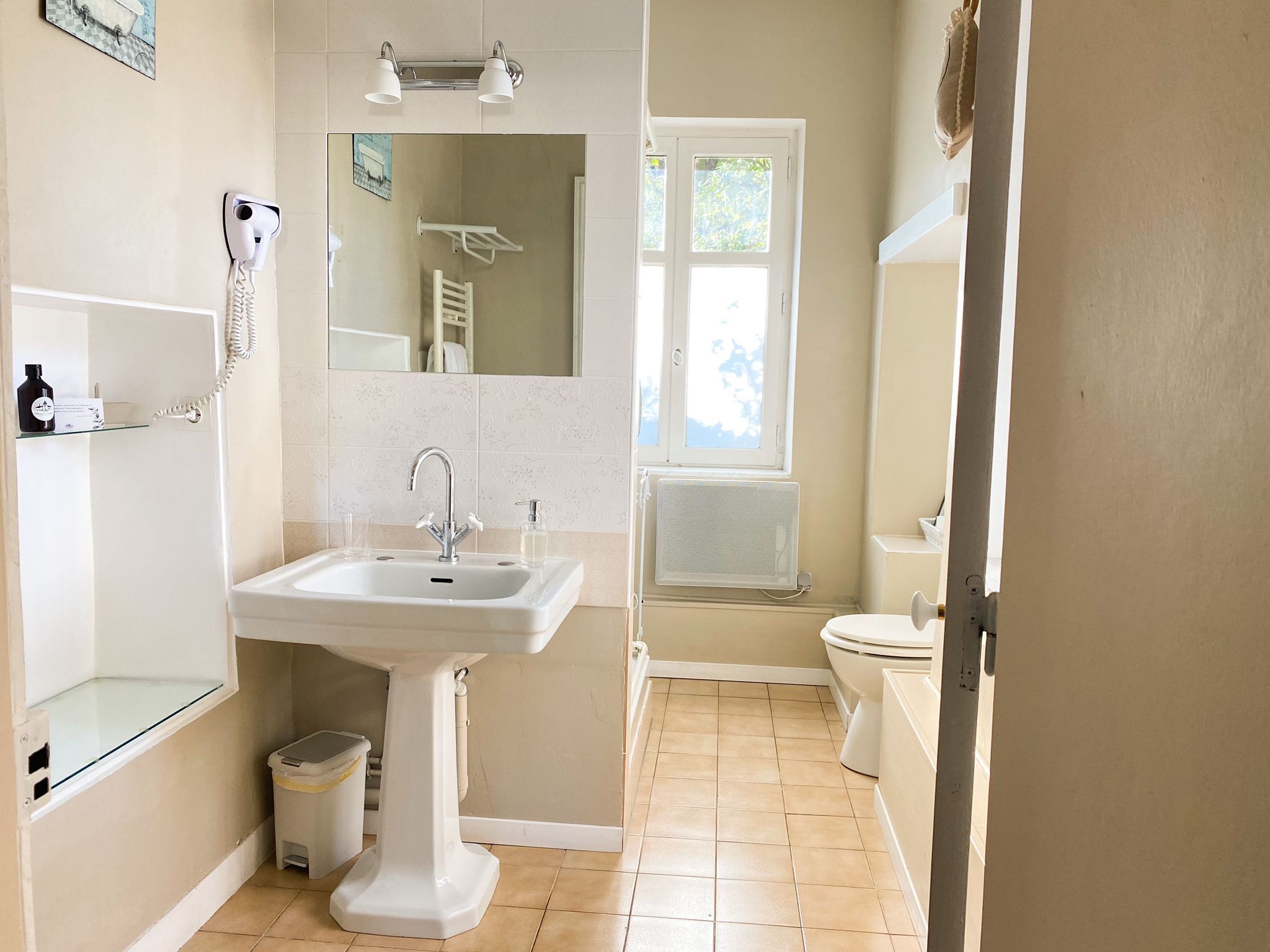 A bathroom with a pedestal sink, a handheld hair dryer, a towel rack, a toilet, and a window providing natural light.