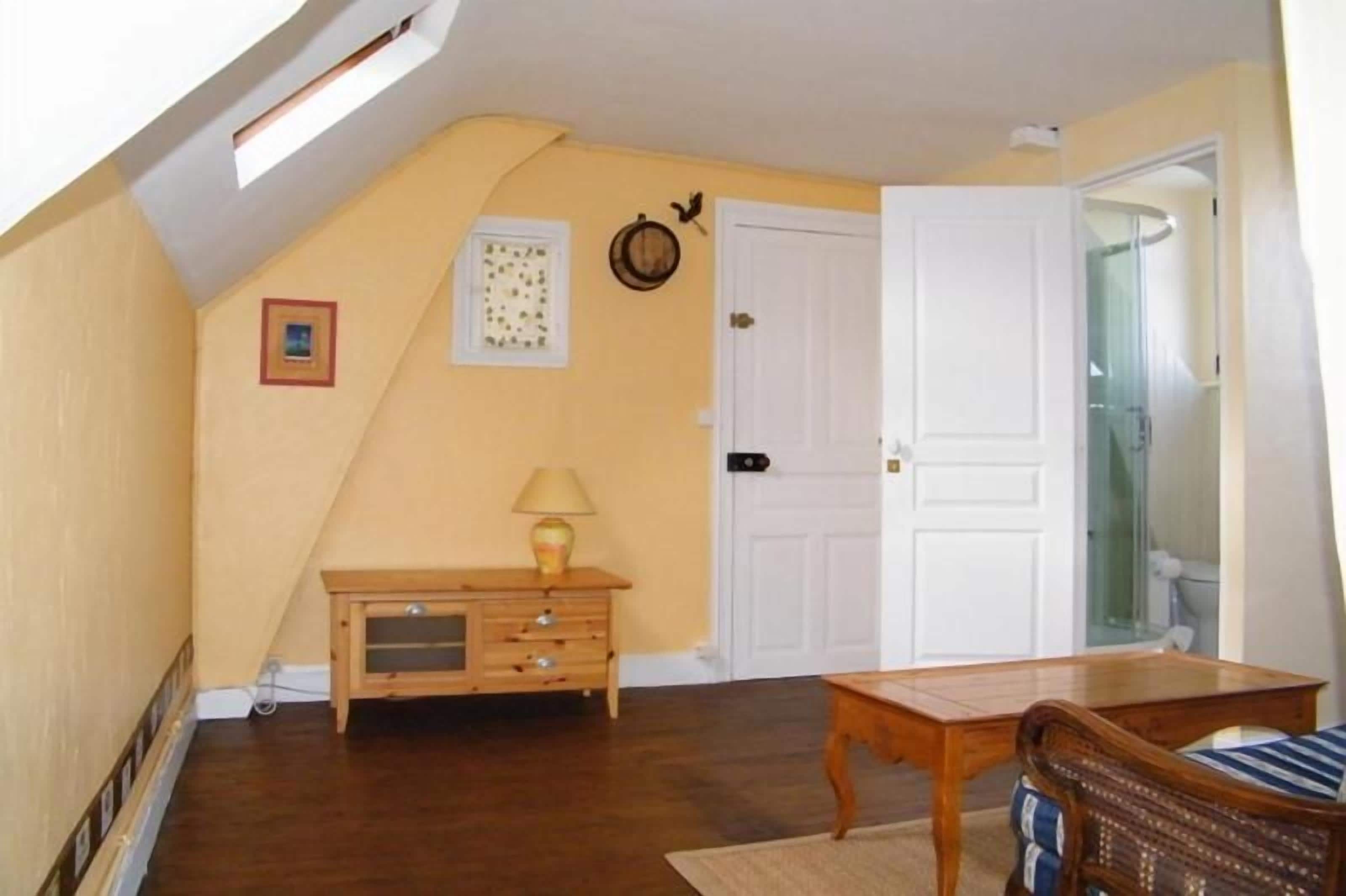 Una habitación con techo inclinado, un aparador de madera, una mesa, una silla y una puerta que da al baño.