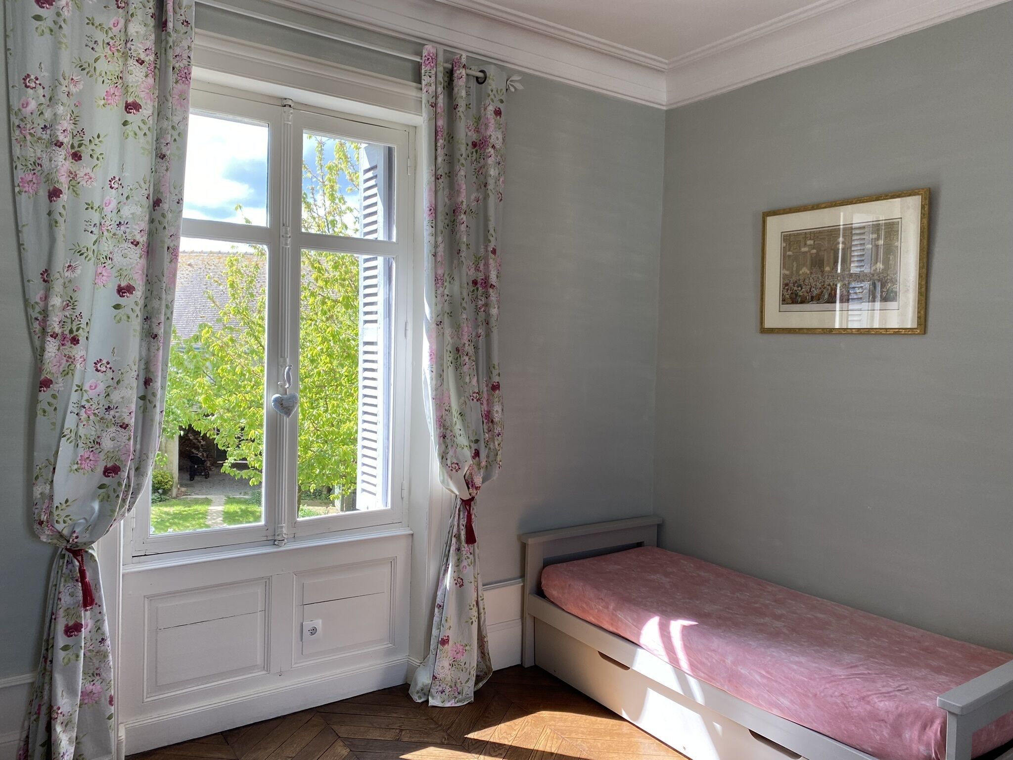 Une chambre avec un lit simple, une fenêtre ornée de rideaux à motifs floraux et un tableau encadré accroché au mur.