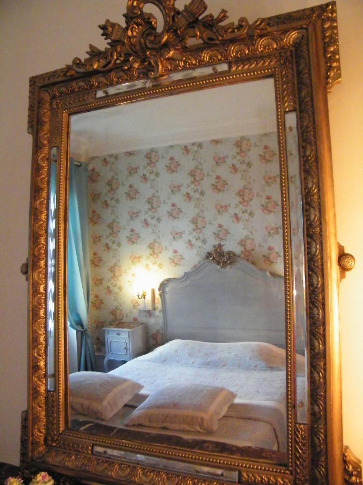Una habitación con un espejo grande y decorativo que refleja una cama con almohadas y una mesita de noche con lámpara.
