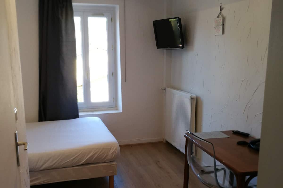 Un petit logement composé d’une seule pièce, comprenant un lit, un bureau, une chaise, un téléviseur fixé au mur et une fenêtre avec des rideaux.
