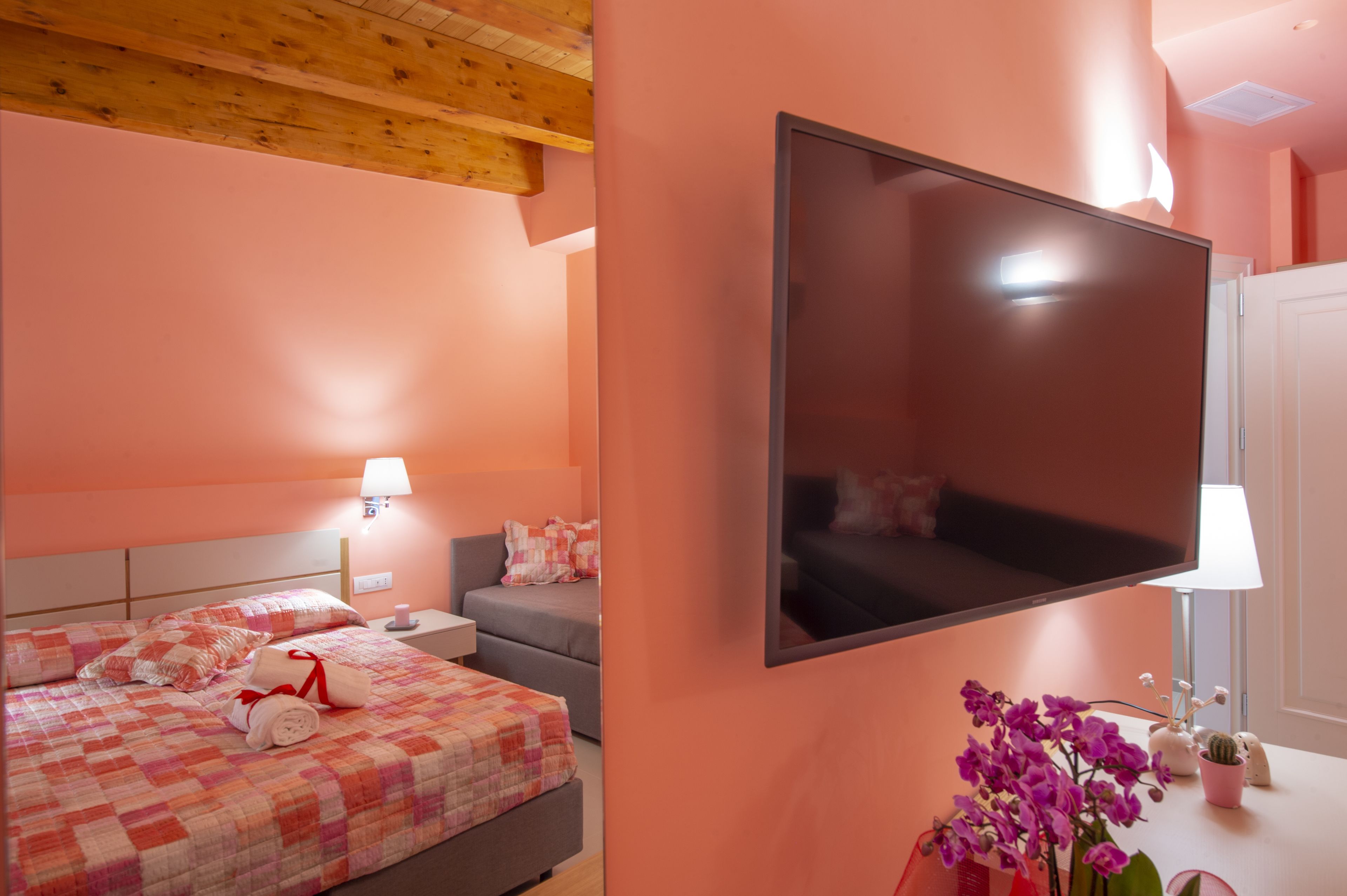 A hotel room with a bed, a TV mounted on the wall, a nightstand, a lamp, and a small table with a plant.