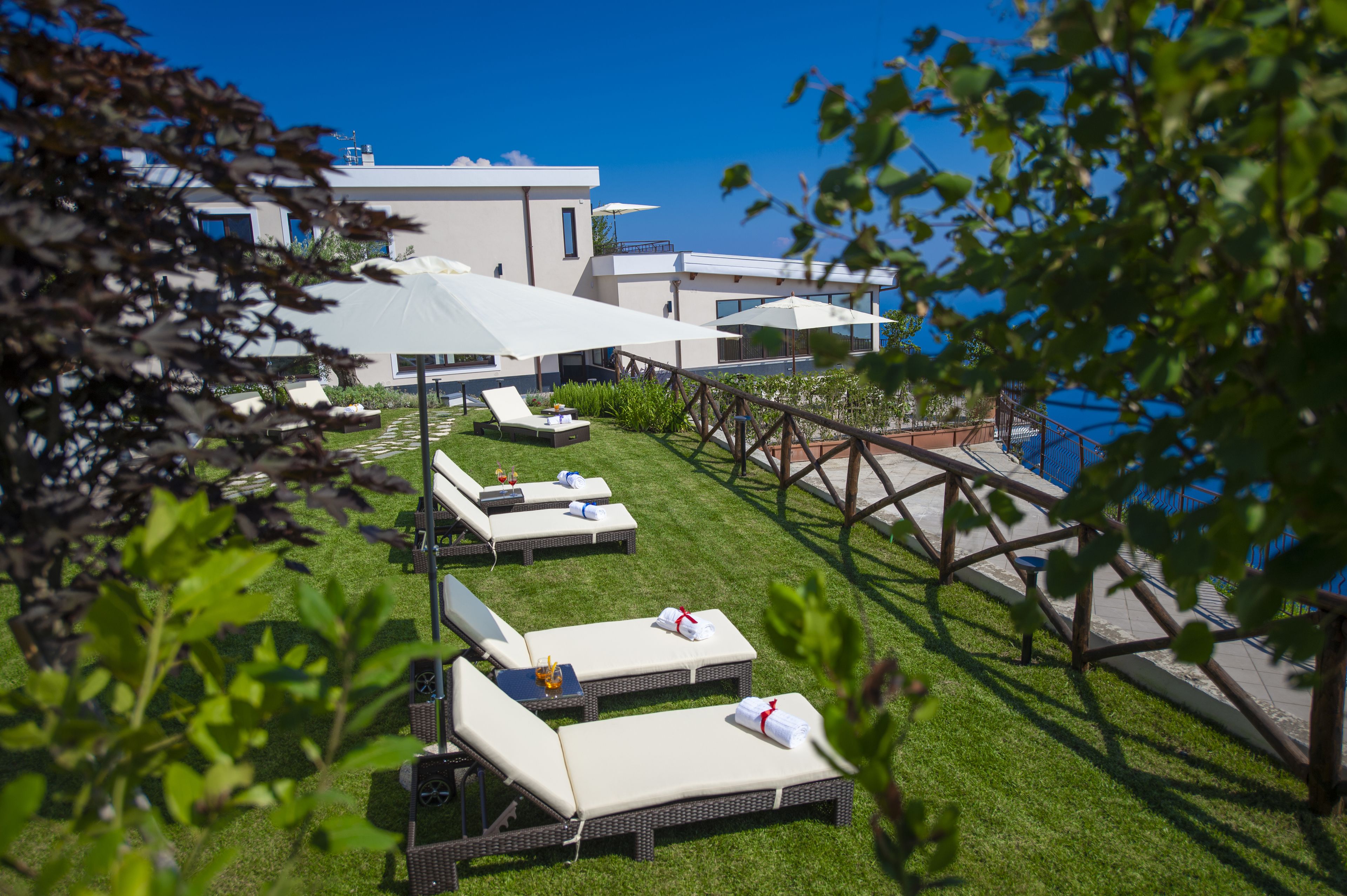 A modern outdoor lounge area with sunbeds and umbrellas, a wooden deck, and a view of a building with a blue sky.