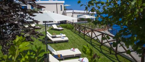 A modern outdoor lounge area with sunbeds and umbrellas, a wooden deck, and a view of a building with a blue sky.
