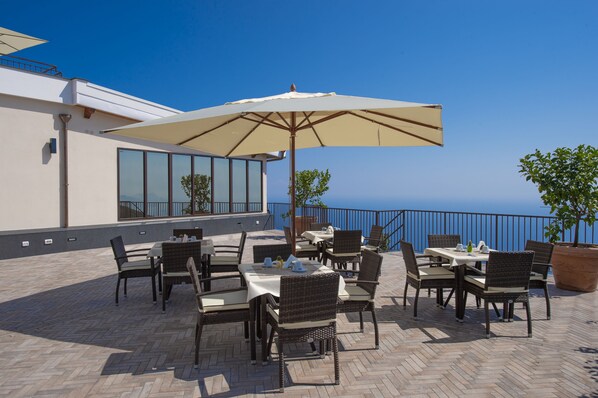 An outdoor patio with tables and chairs, a large umbrella, and a view of the sea.