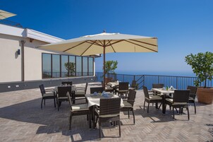 An outdoor patio with tables and chairs, a large umbrella, and a view of the sea.