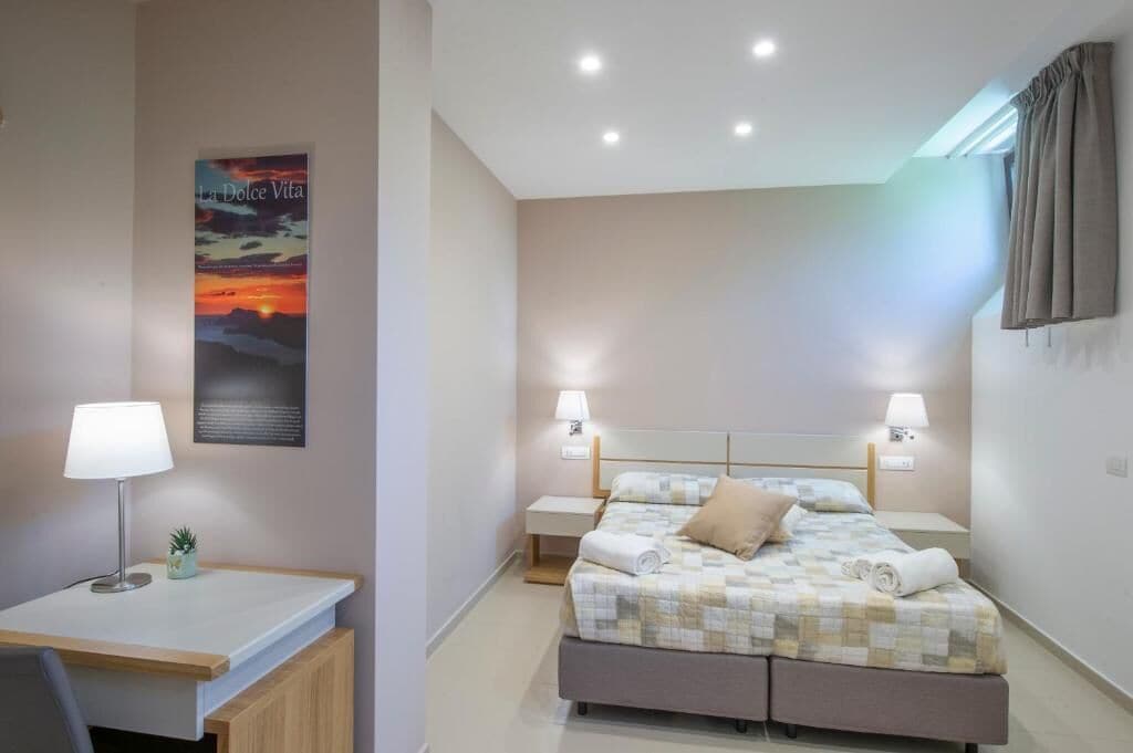 A bedroom with a bed, two bedside lamps, a desk with a lamp, a small plant, and a poster on the wall.