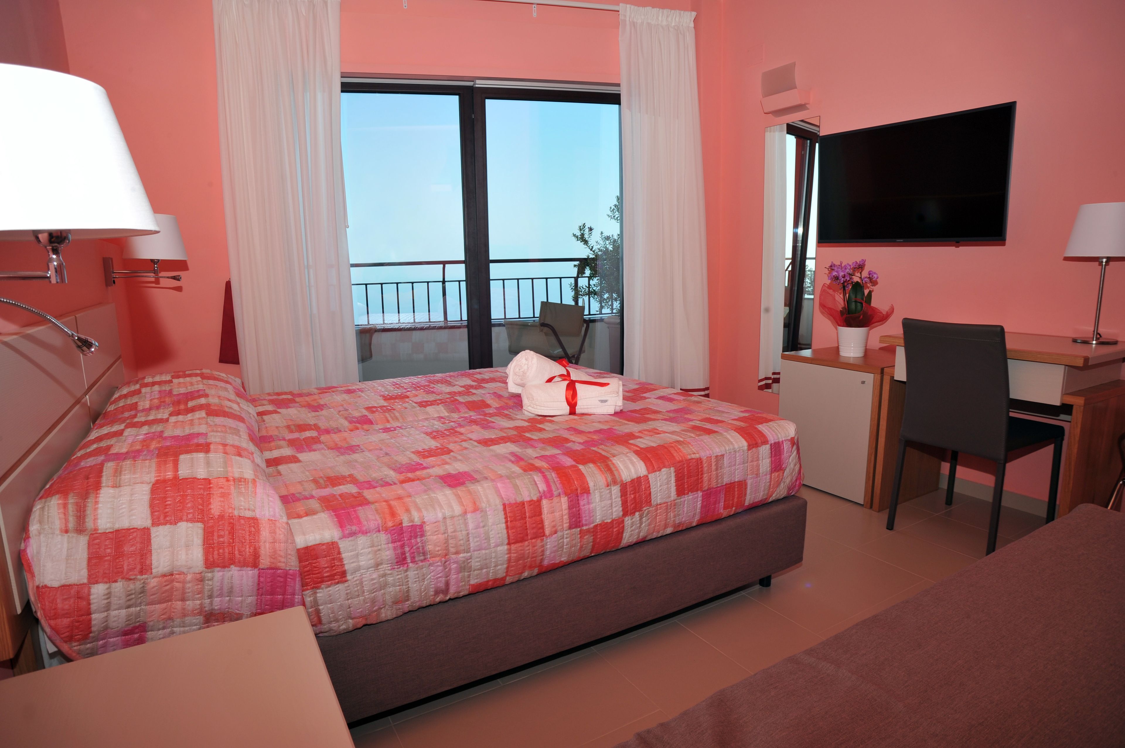 A hotel room with a bed, a desk, a chair, a TV, and a balcony with a chair.