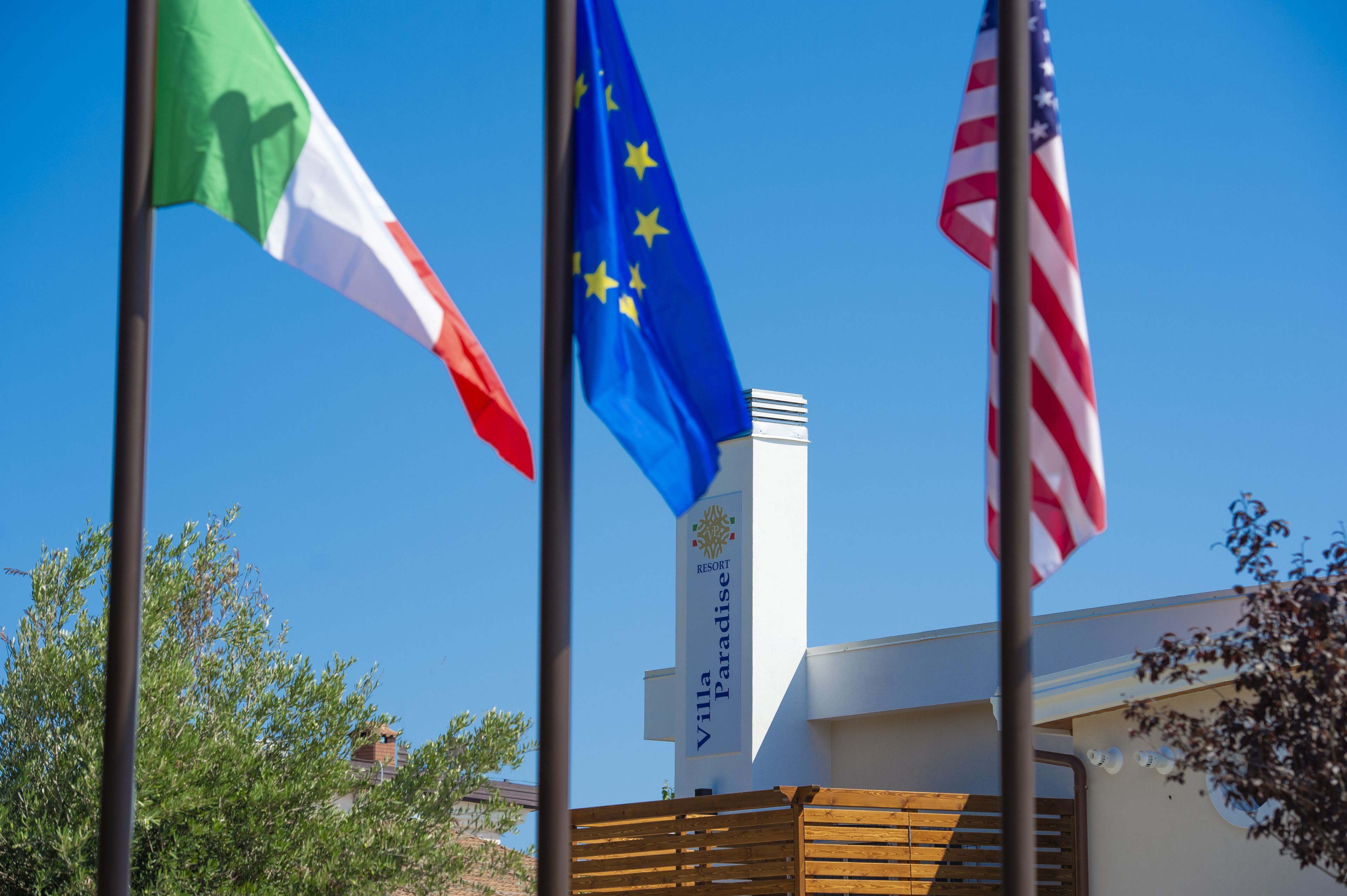 Three flags on flagpoles in front of a building with a sign that reads "Villa Paradis Resort".