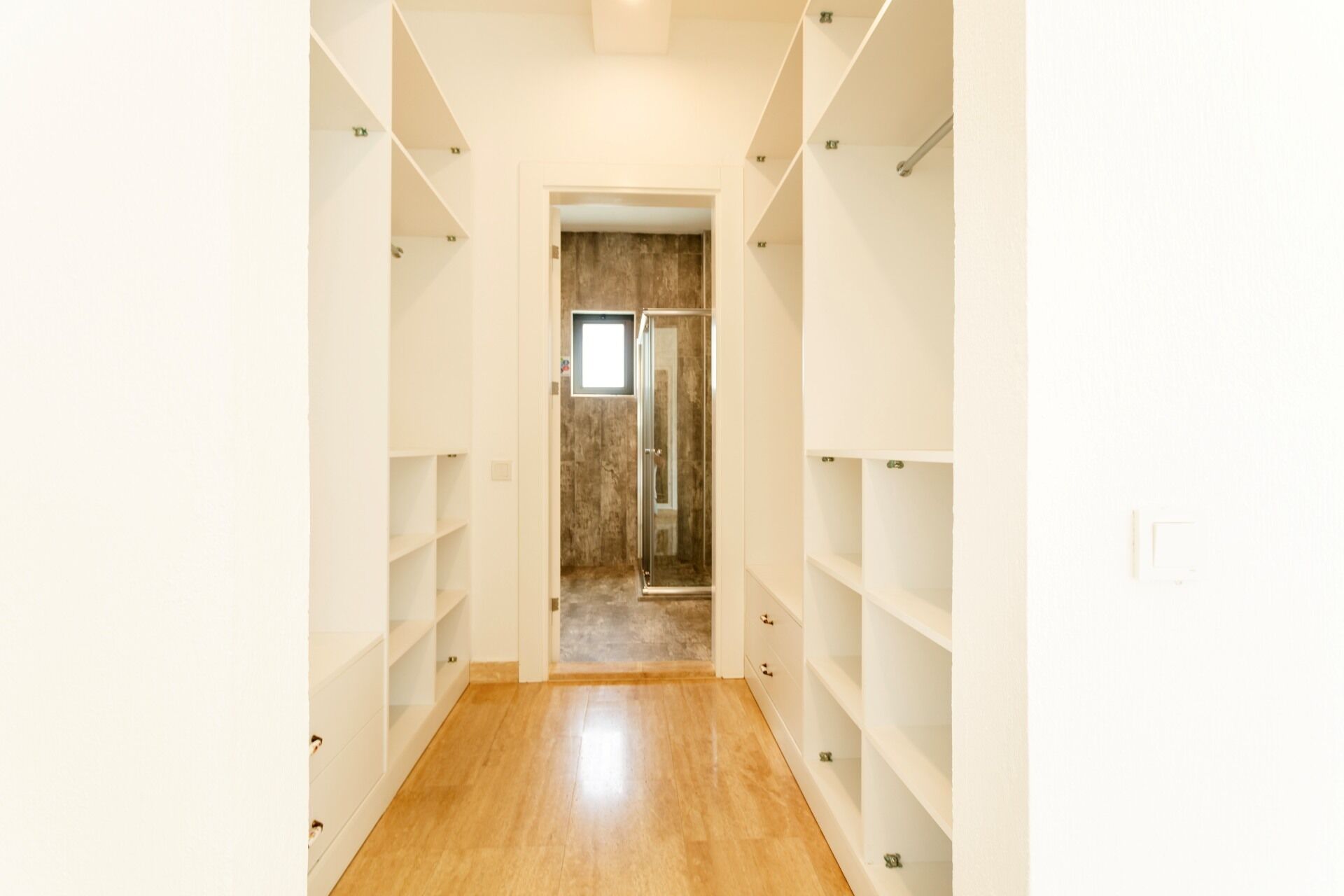A narrow hallway with wooden flooring leading to a room with a glass door. Shelves line the walls on both sides.