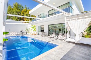 Ein moderner Poolbereich im Freien mit Liegestühlen und einem Esstisch, umgeben von Glaswänden und Blick auf Bäume.