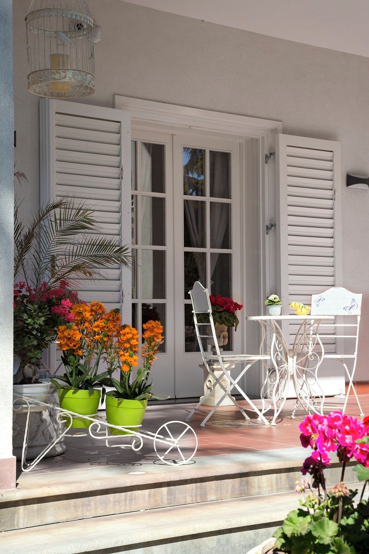 Un patio con contraventanas blancas, una mesa y sillas de metal, y macetas con flores naranjas.
