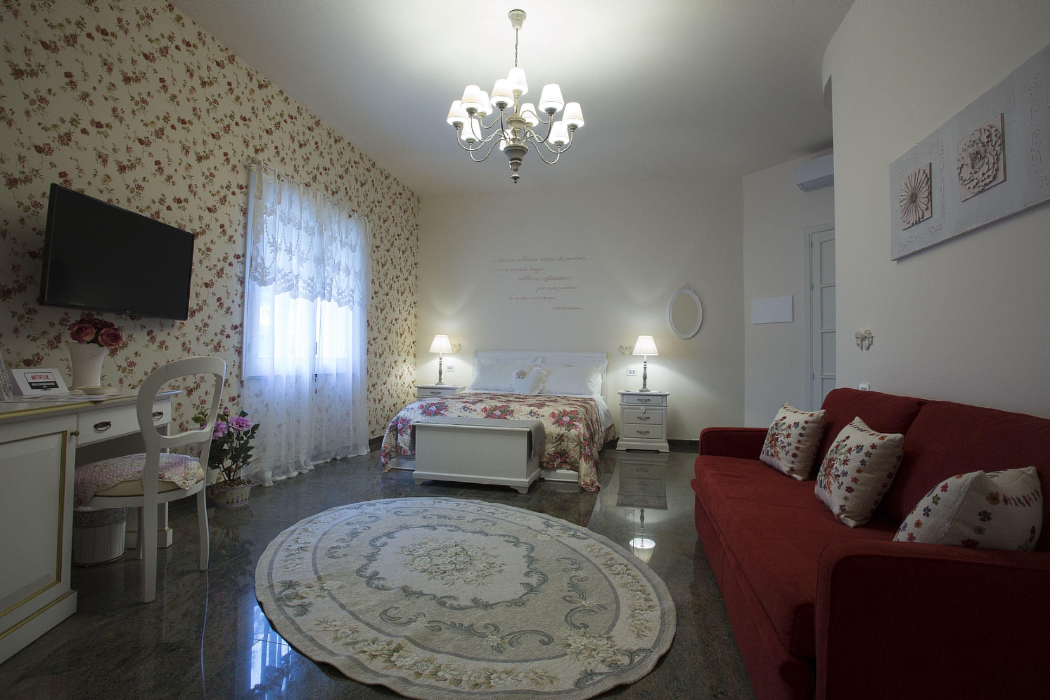 A bedroom with a bed, bedside tables, a red sofa, a round rug, a TV, and a chandelier.
