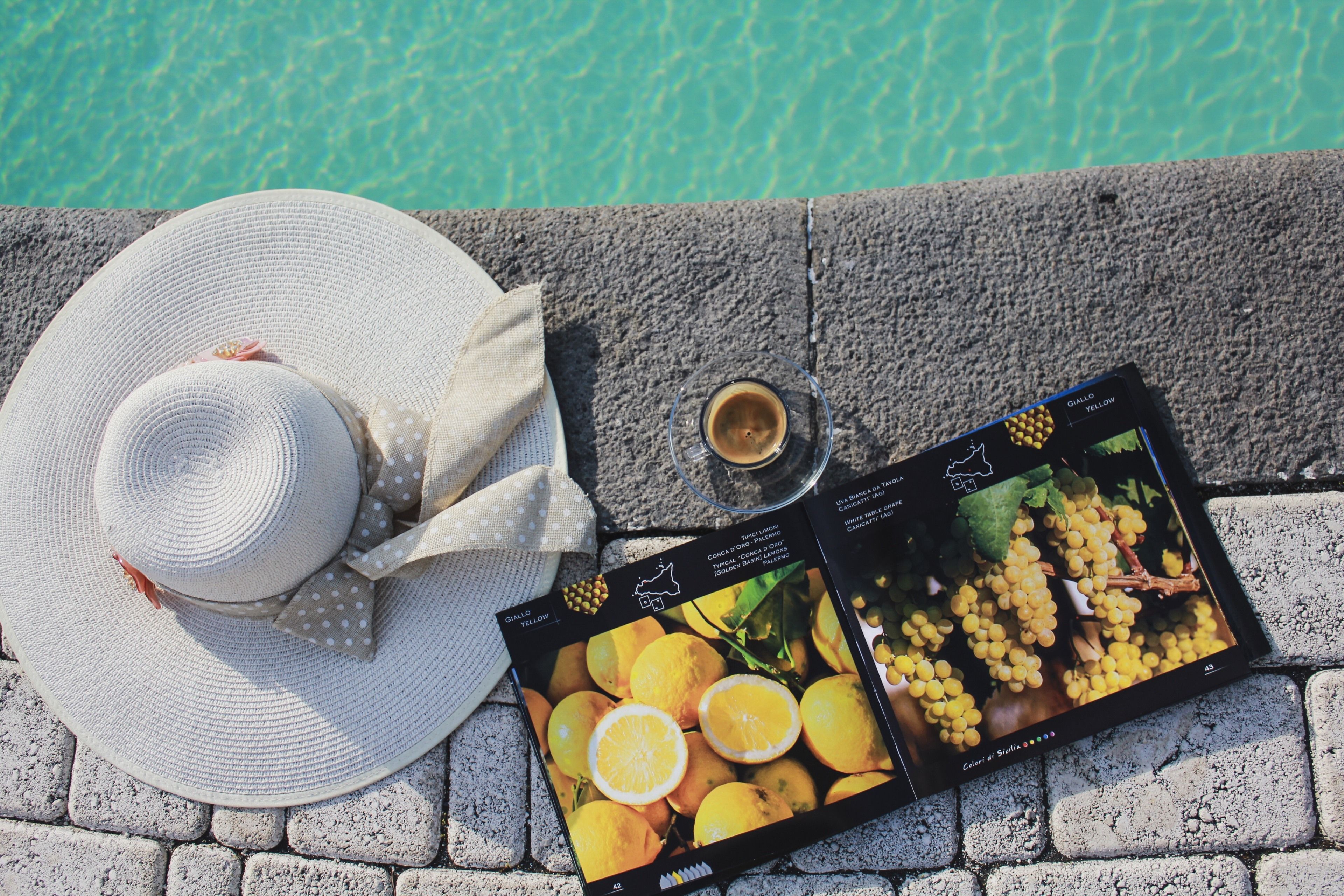 A white straw hat, a glass of drink, and two wine bottle labels on a stone surface near a pool.