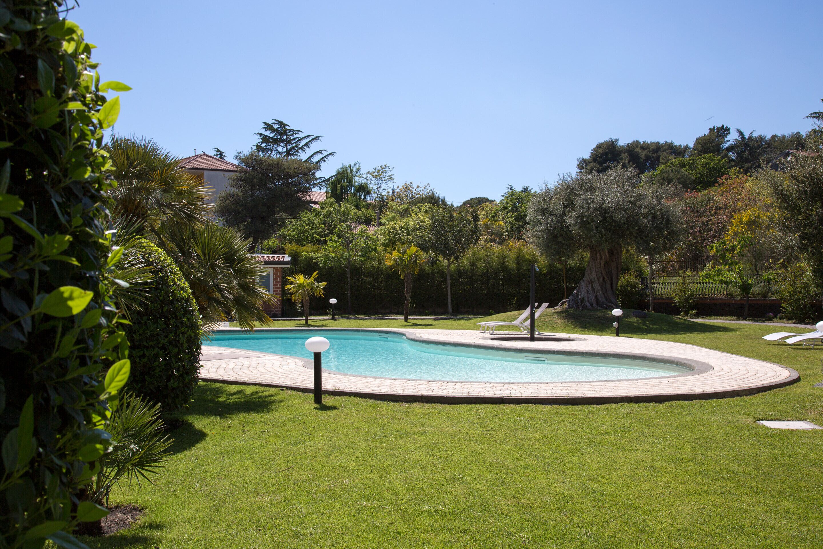 Una piscina con un área verde alrededor, algunas sillas de descanso y un edificio al fondo.
