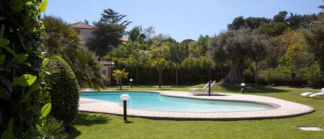 Una piscina con un área verde alrededor, algunas sillas de descanso y un edificio al fondo.