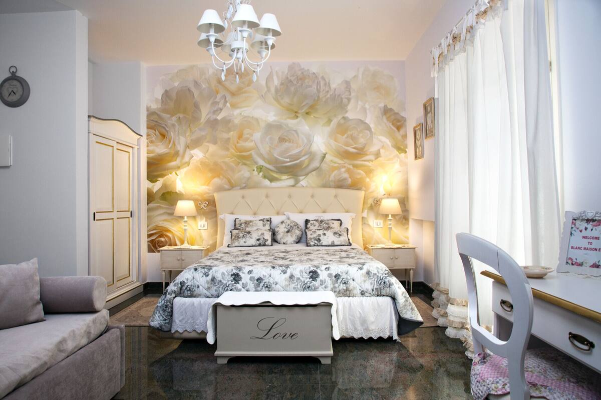 Une chambre avec un grand décor mural floral sur le mur, un lit avec un banc orné du mot « Amour », une chaise blanche et une table de chevet avec une lampe.