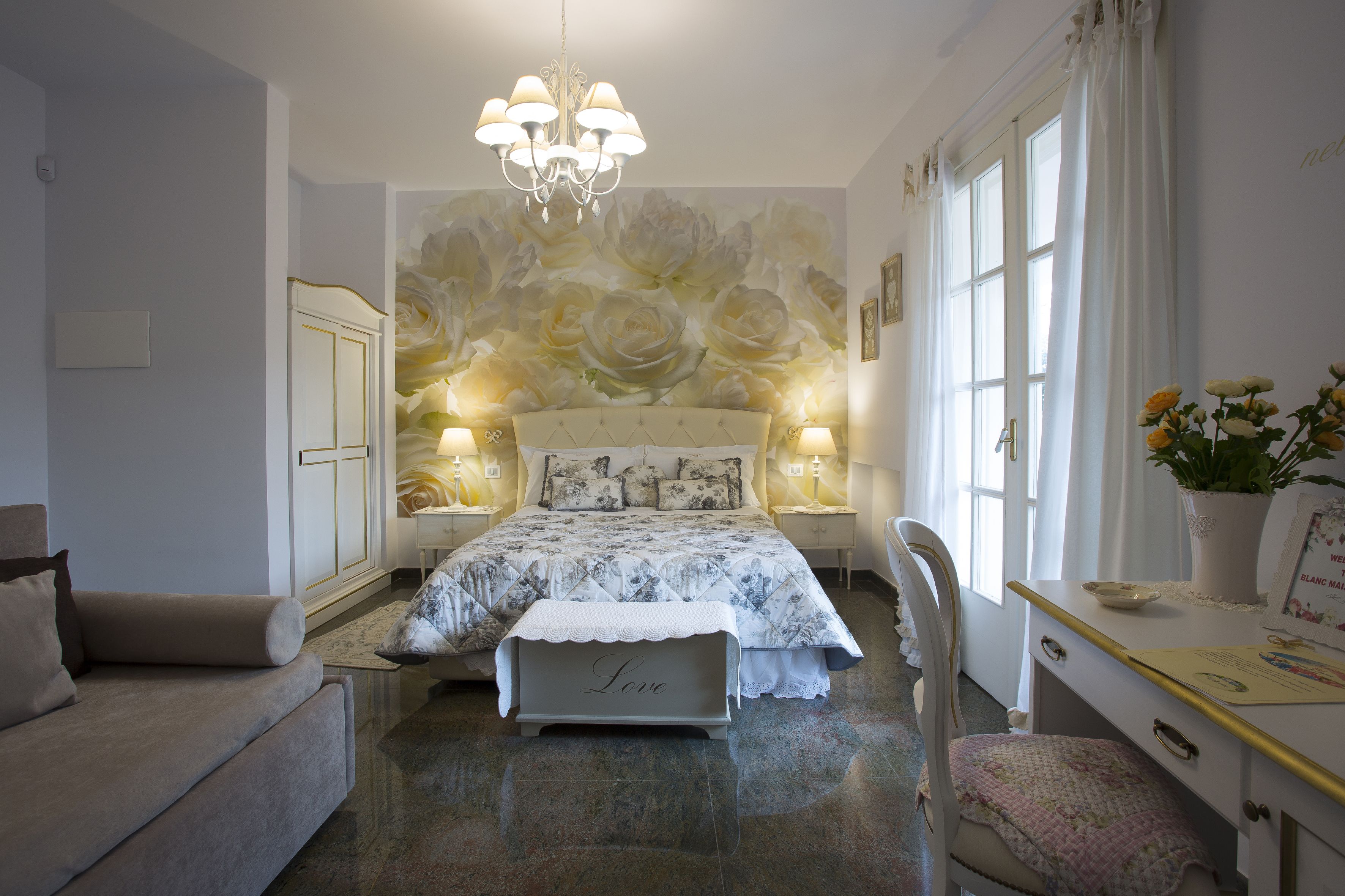 A bedroom with a large bed, two bedside tables with lamps, a chair, a desk with a vase of flowers, and a chandelier.