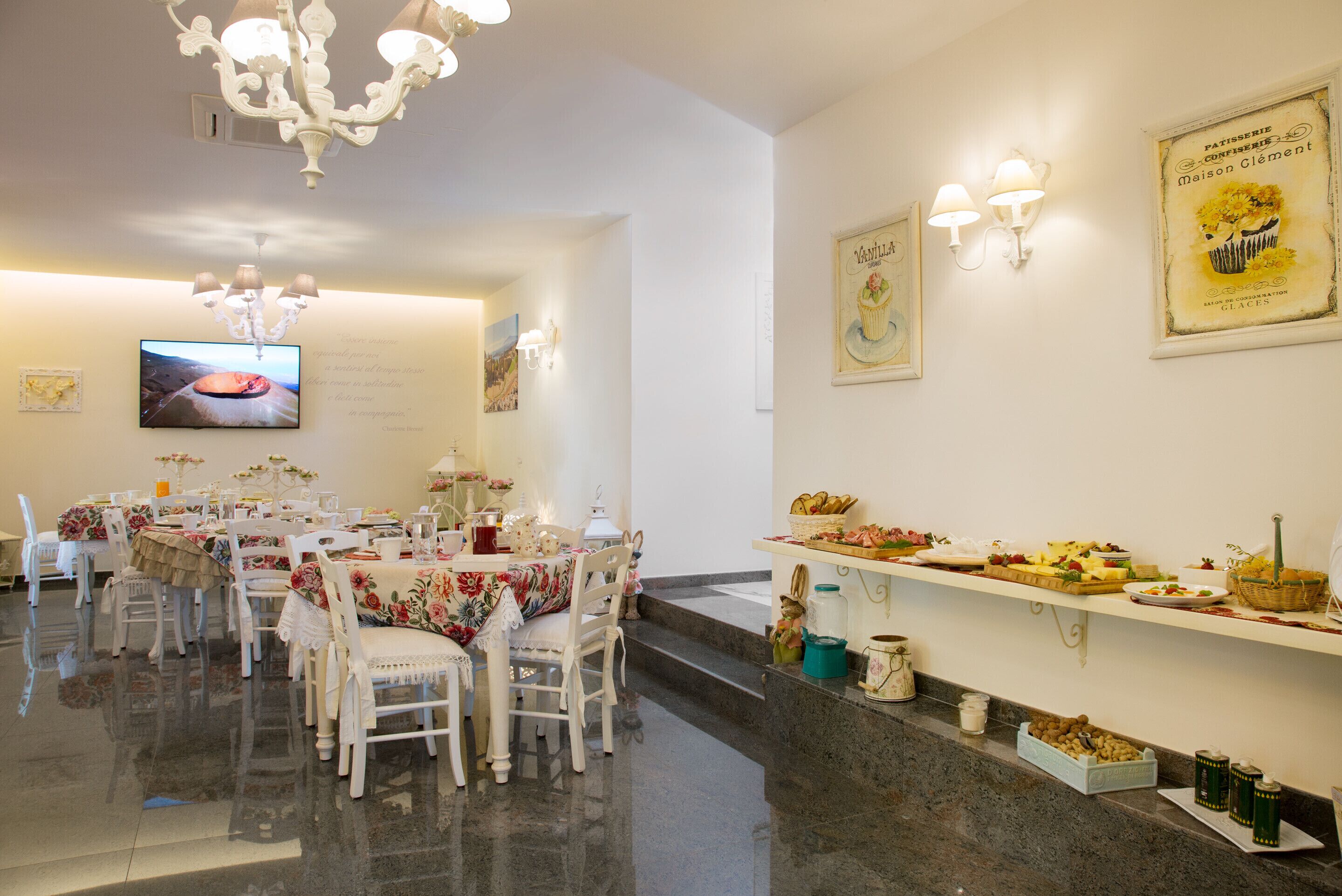 A dining area with white tables and chairs, a chandelier, wall-mounted lights, and a buffet with various dishes.