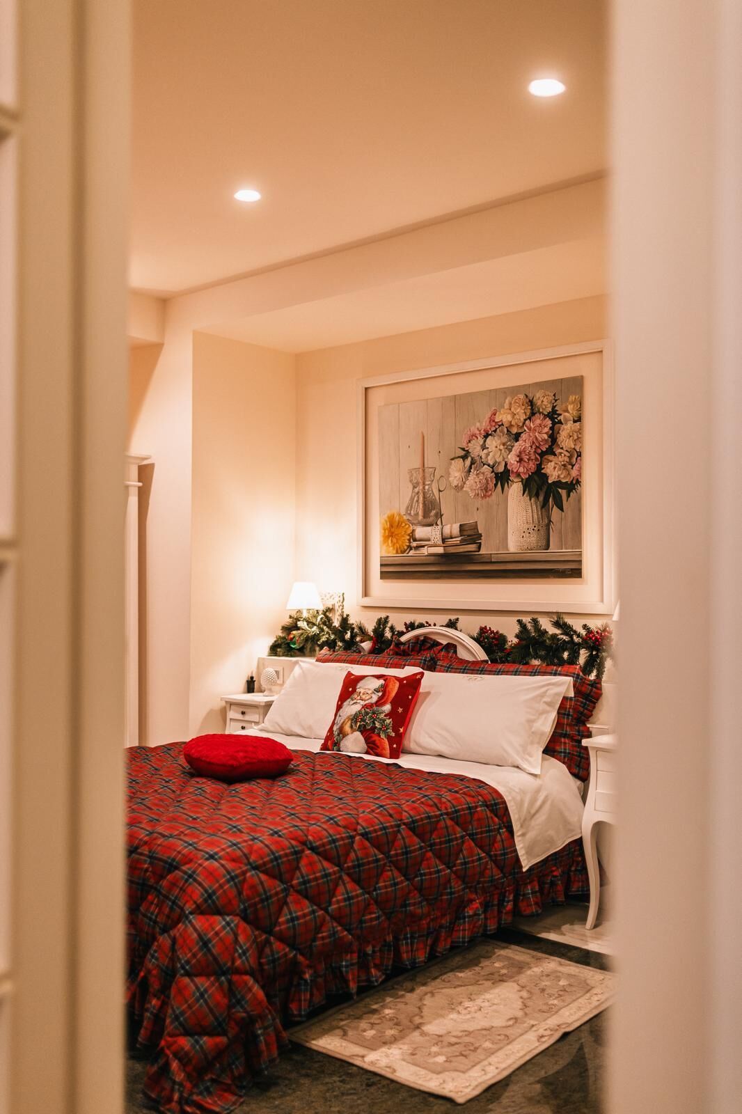 Un dormitorio con una cama cubierta con una colcha a cuadros rojos, una obra de arte floral enmarcada sobre la cama y una mesita de noche con una lámpara.