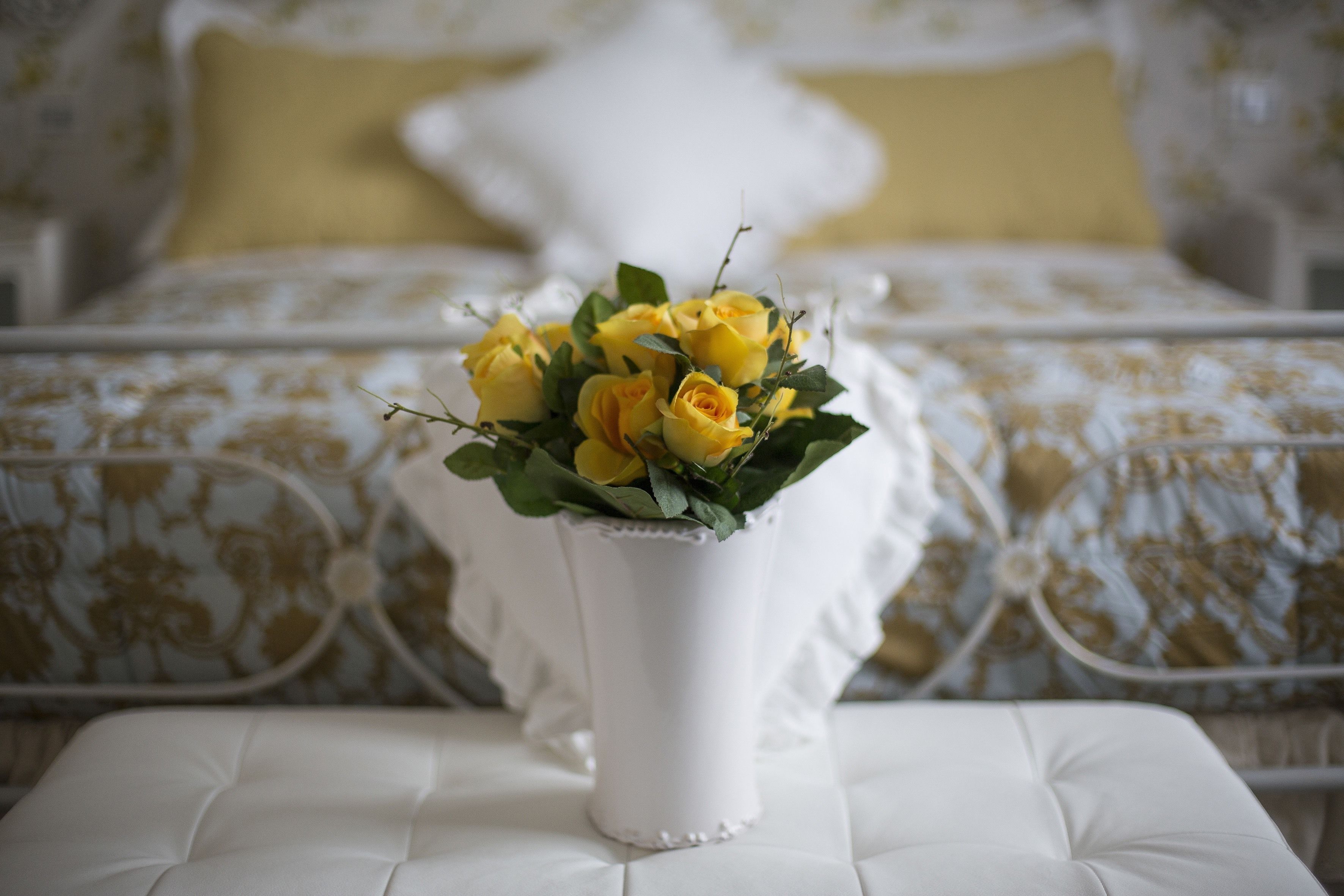 Un jarrón blanco con rosas amarillas sobre una mesa blanca con cojines, en una habitación con cama y ropa de cama estampada.
