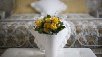 Un jarrón blanco con rosas amarillas sobre una mesa blanca con cojines, en una habitación con cama y ropa de cama estampada.