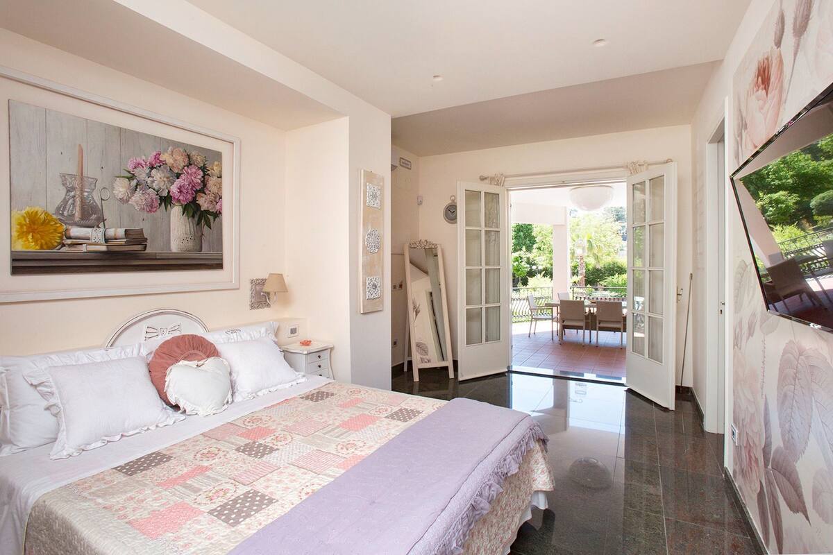 Une chambre à coucher comprenant un lit, une table de chevet, une commode et un tableau représentant des fleurs. Une porte donne accès à un balcon aménagé avec du mobilier d’extérieur.