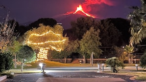 Un espace patio avec une table et des chaises, un arbre décoré de guirlandes lumineuses et, au loin, une montagne sur laquelle se dresse un volcan.