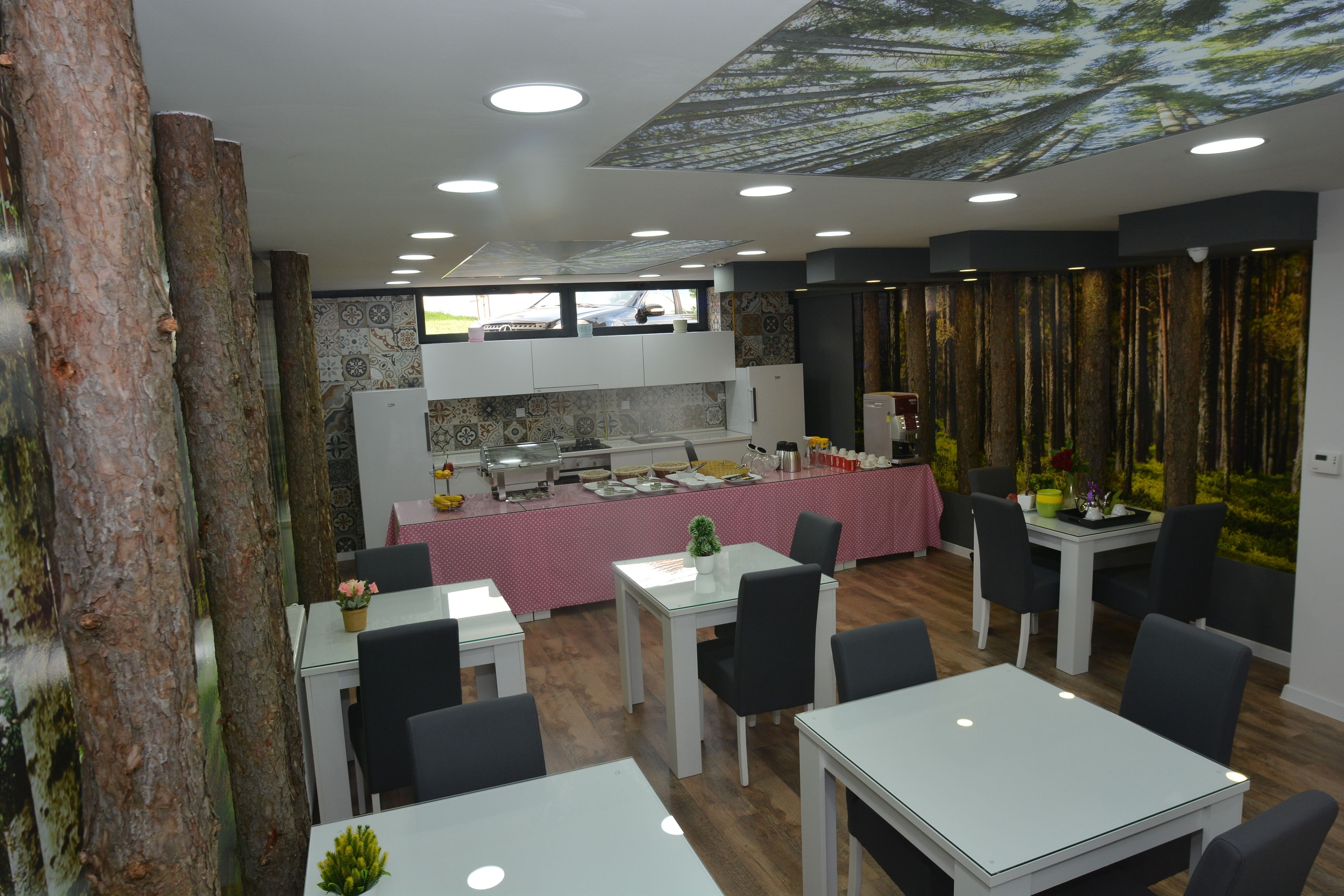 Un restaurant au décor moderne, avec une salle à manger centrale dotée de tables blanches et de chaises noires, un espace cuisine avec une nappe rose et un plafond décoré sur le thème de la forêt.