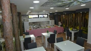Un restaurant au décor moderne, avec une salle à manger centrale dotée de tables blanches et de chaises noires, un espace cuisine avec une nappe rose et un plafond décoré sur le thème de la forêt.
