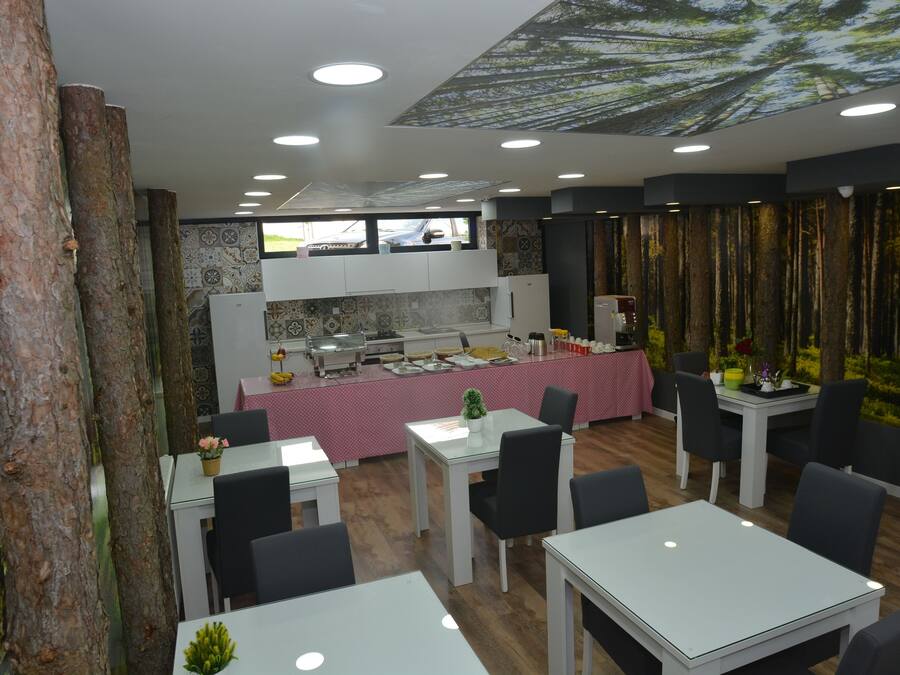 Un restaurant au décor moderne, avec une salle à manger centrale dotée de tables blanches et de chaises noires, un espace cuisine avec une nappe rose et un plafond décoré sur le thème de la forêt.