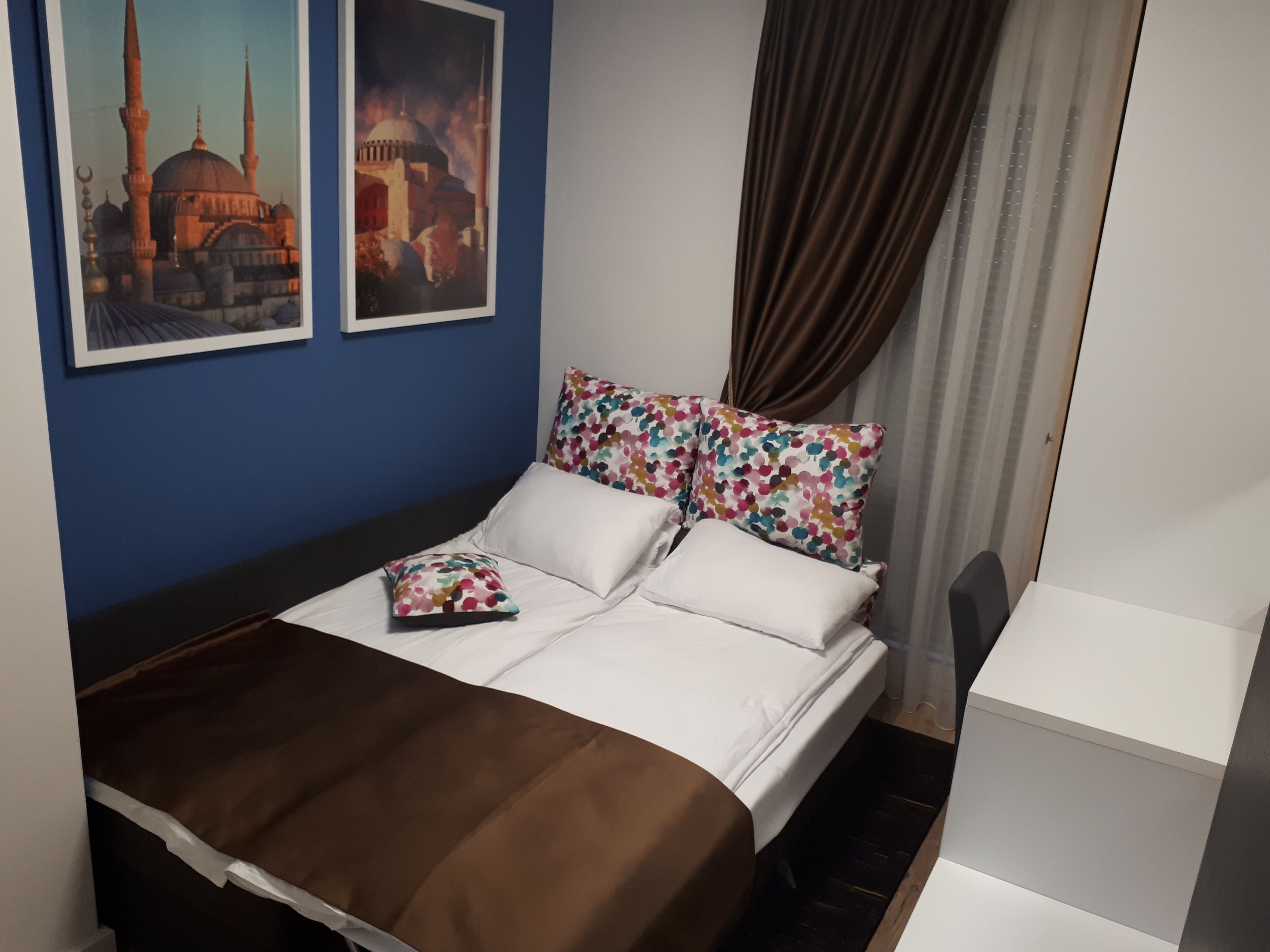 A neatly made bed with white and brown bedding, a colorful floral pillow, and a framed picture of a building on the wall.