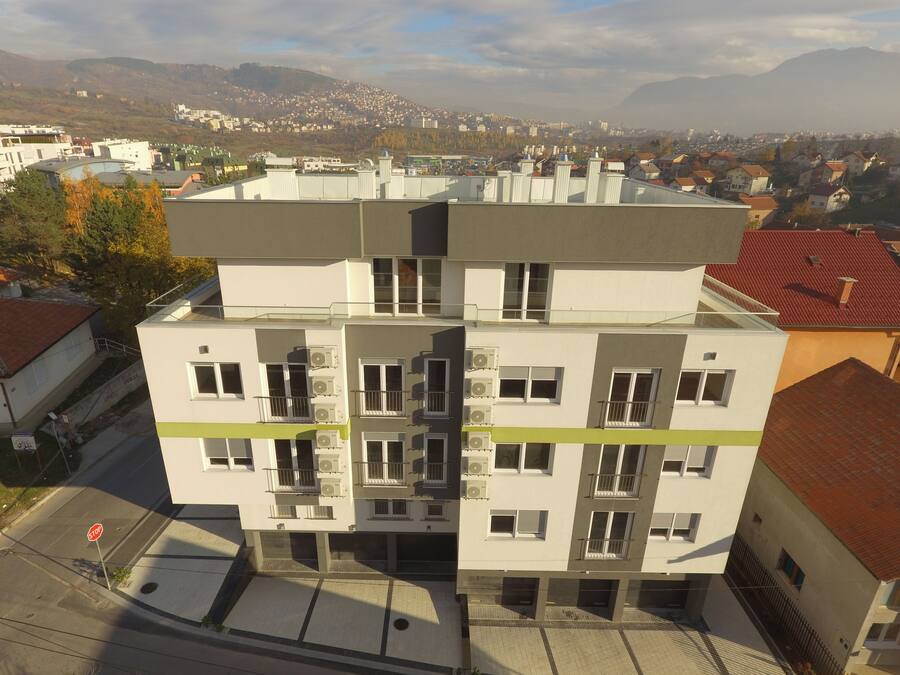 A multi-story building with balconies and air conditioning units, surrounded by a residential area with houses and mountains in the background.