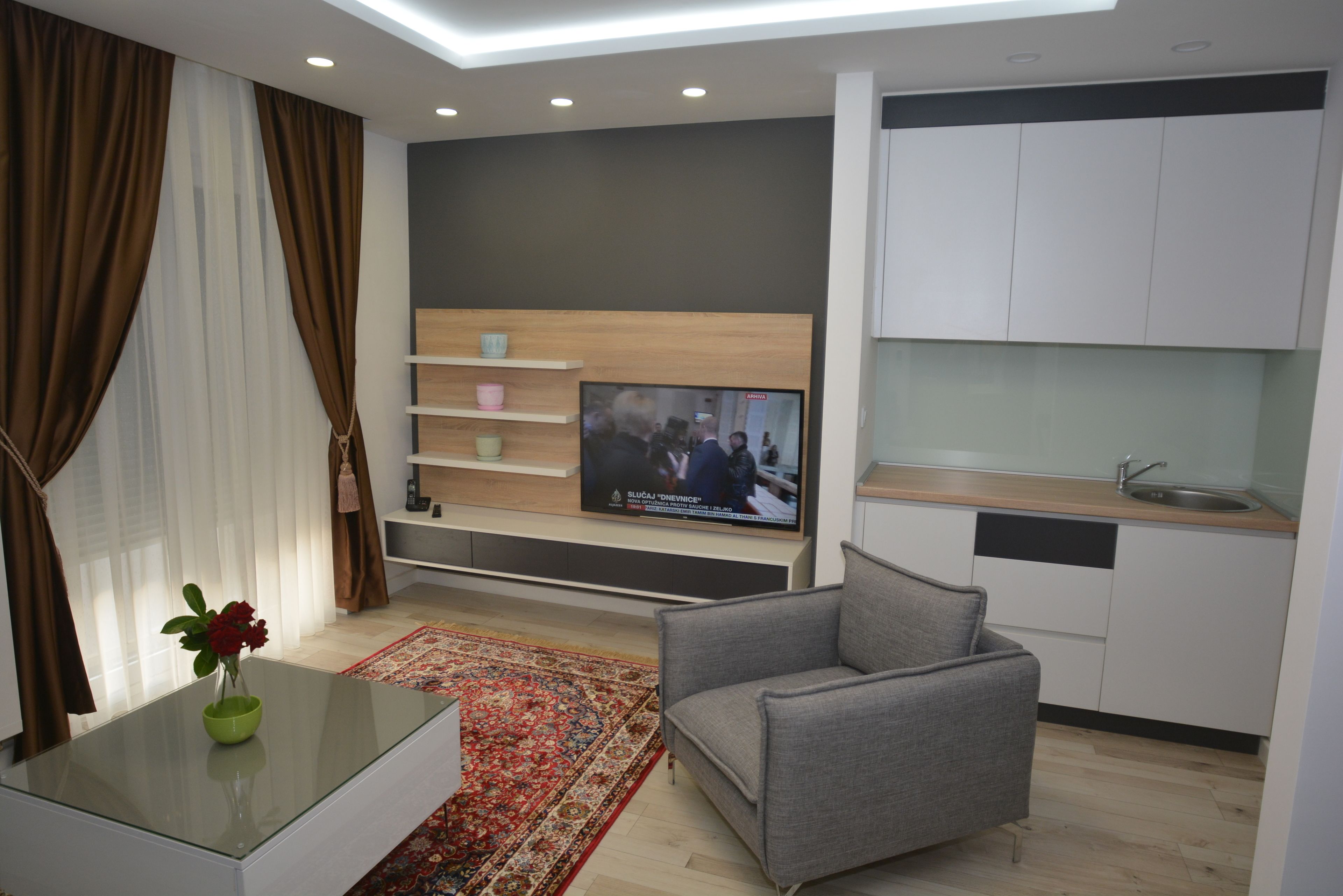 Uma sala de estar moderna com televisão de tela plana, um sofá cinza, uma mesa de centro de vidro com um vaso e uma área de cozinha com armários brancos e bancada de madeira.