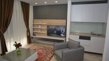 Uma sala de estar moderna com televisão de tela plana, um sofá cinza, uma mesa de centro de vidro com um vaso e uma área de cozinha com armários brancos e bancada de madeira.