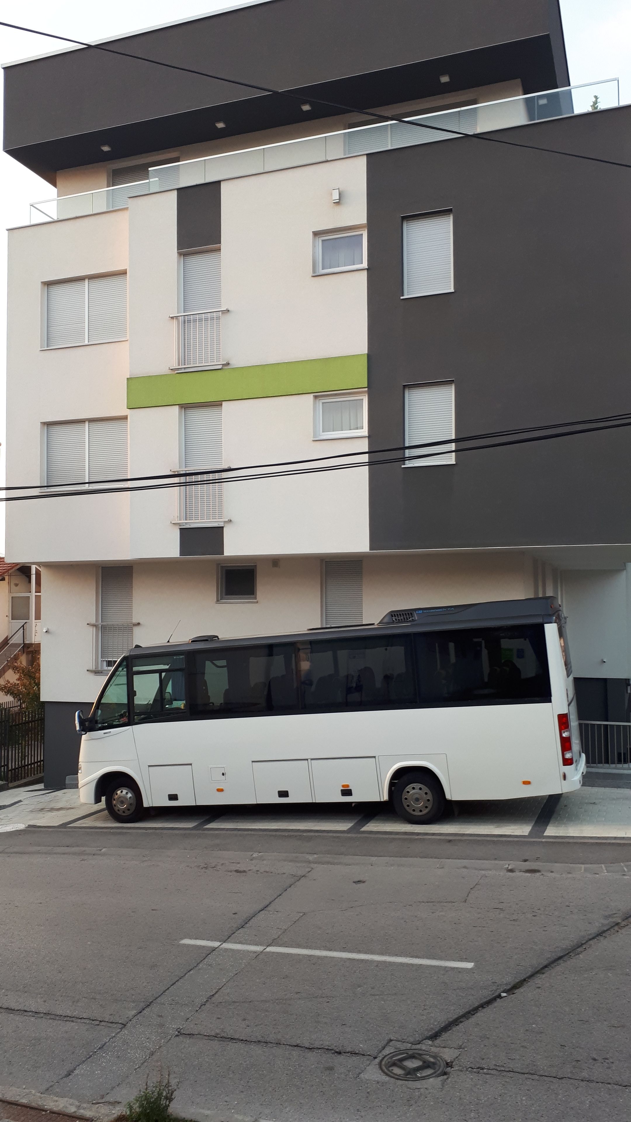 A modern multi-story building with a white and grey facade, a green stripe, and a parked white bus in front.