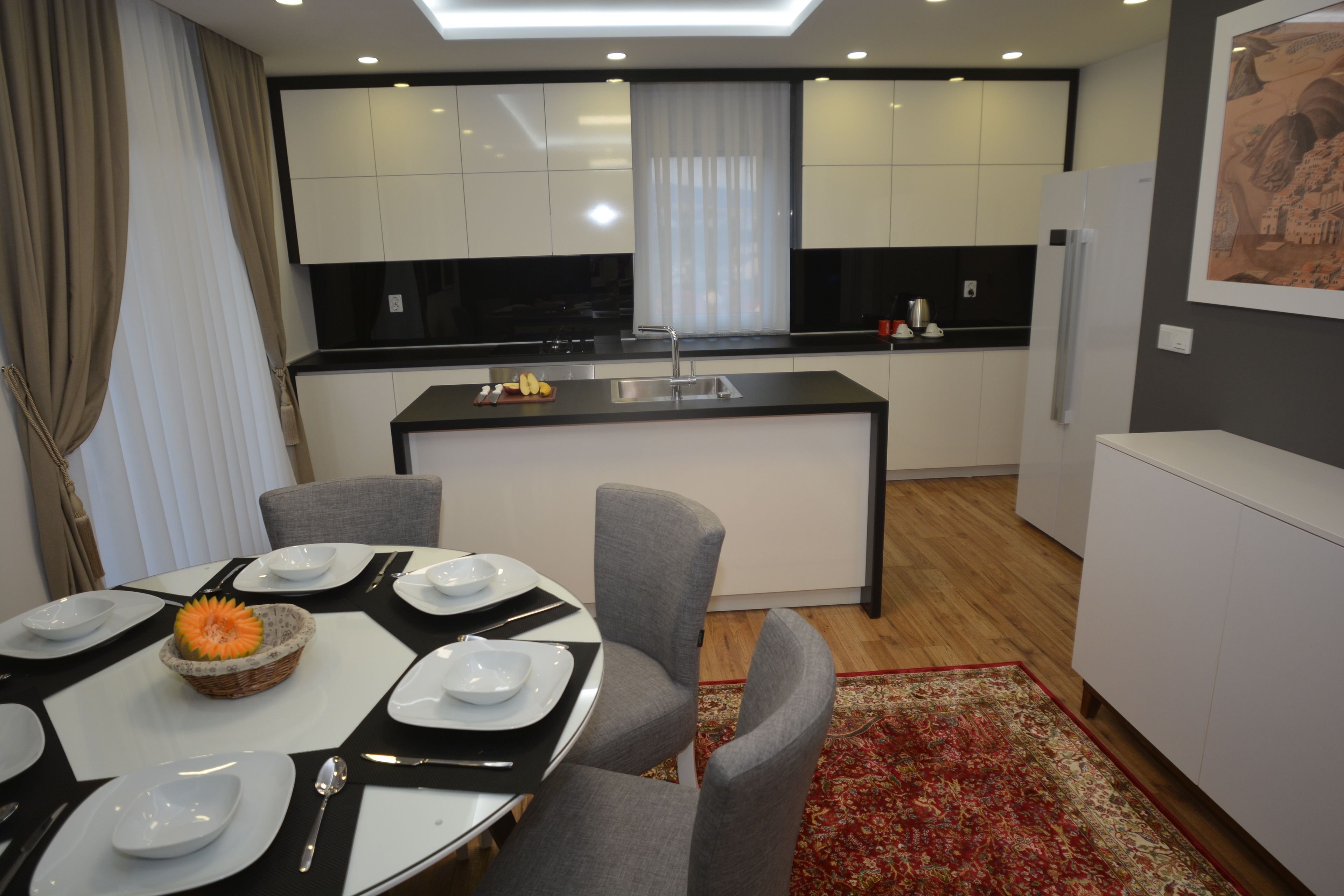 A modern kitchen with white cabinets, a central island, and a dining area with a red rug.